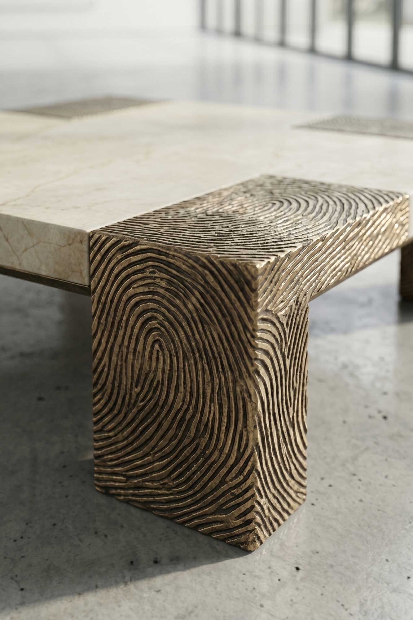 A close-up of a coffee table with a marble top and a gold metal leg featuring a fingerprint pattern. The table sits on a polished concrete floor in a modern, bright room.