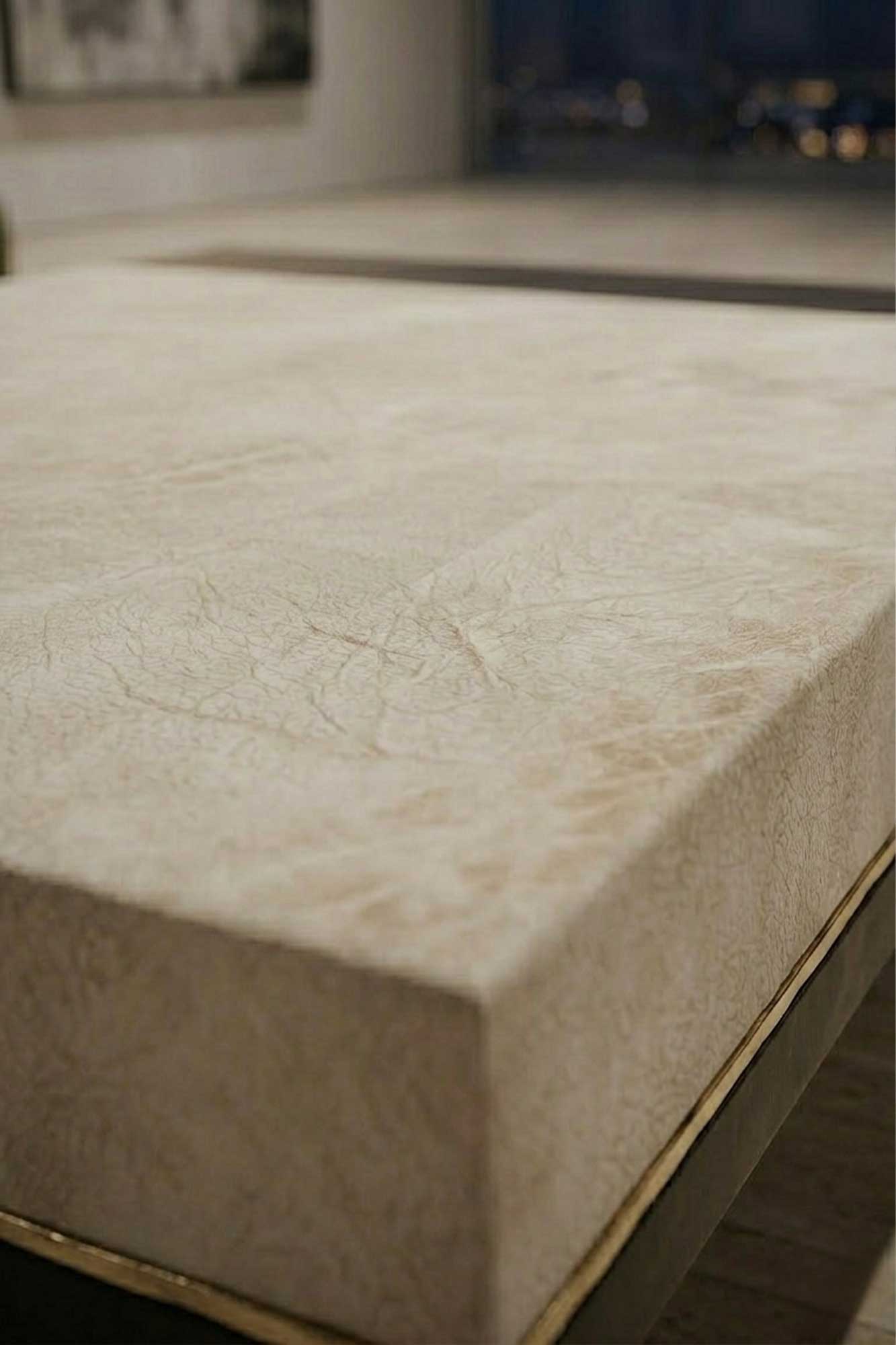 A close-up view of a rectangular, beige tabletop with a subtle textured pattern, placed in a modern indoor setting. The background is slightly out of focus, showing part of another similar surface.