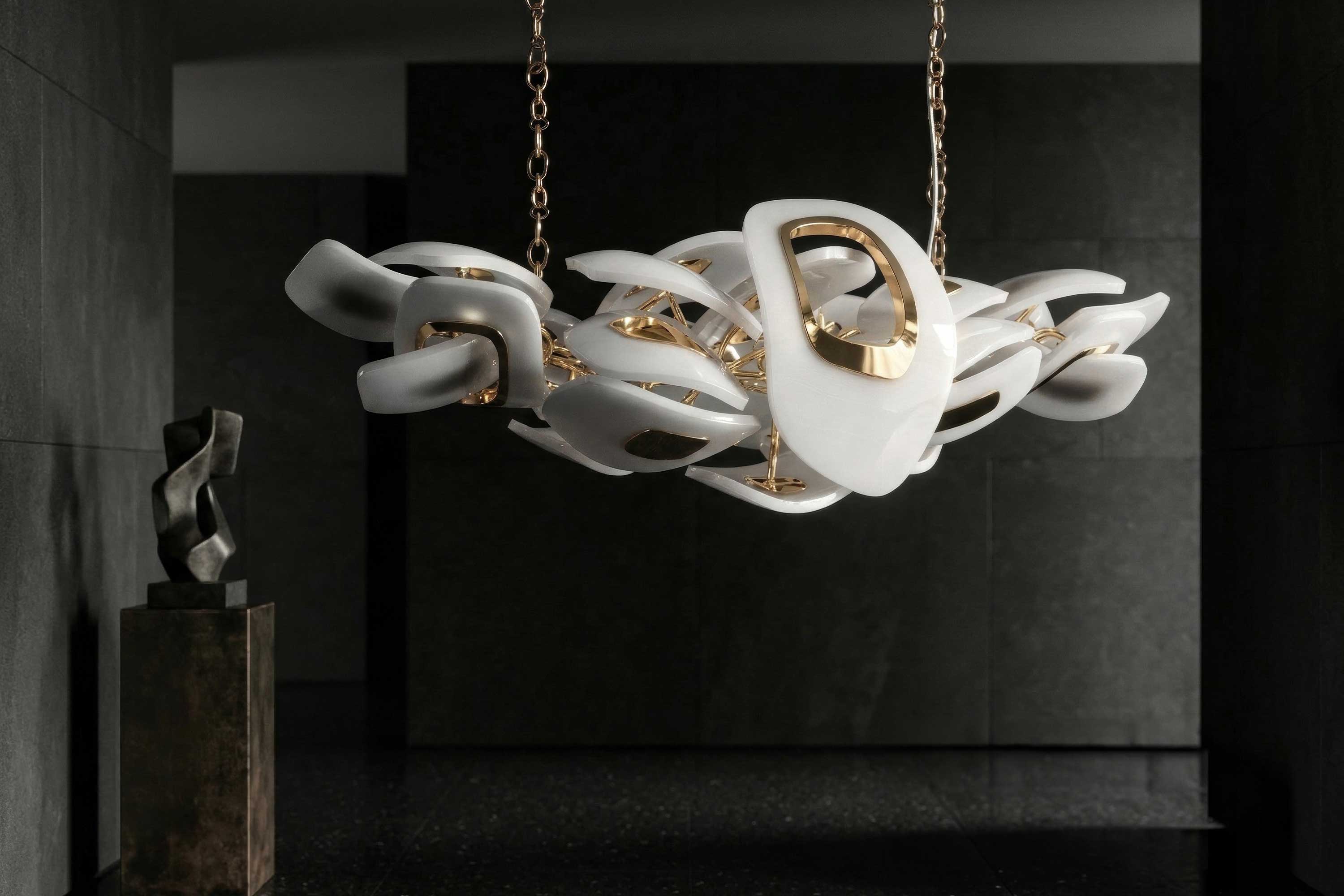 Modern chandelier with interlocking white and gold abstract shapes, suspended by chains in a dark, minimal room. A black abstract sculpture is visible on a pedestal to the left.