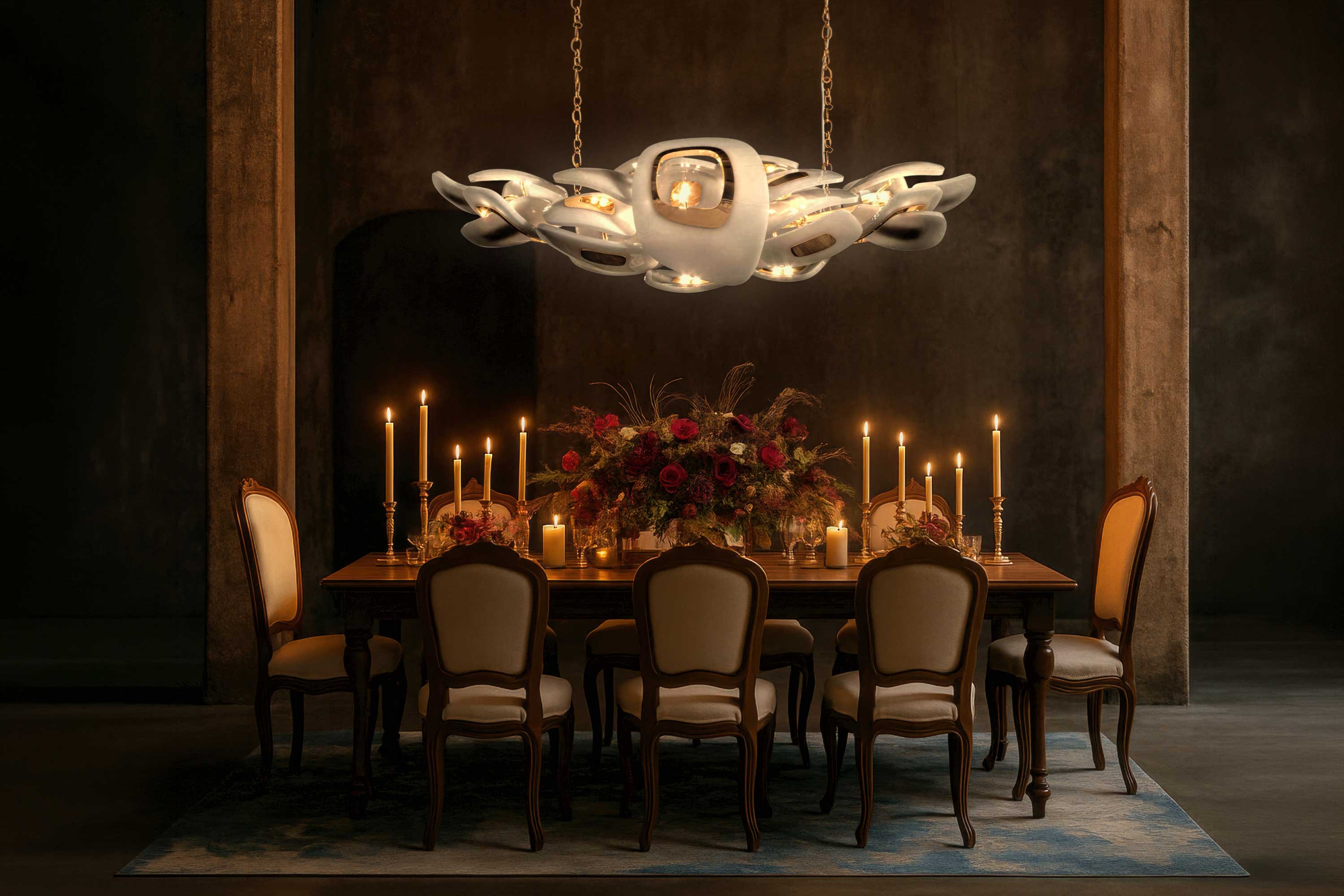 A wooden dining table with eight chairs is set with floral arrangements and lit candles. A large, modern chandelier hangs above. The room is dimly lit with a dark background and concrete pillars.