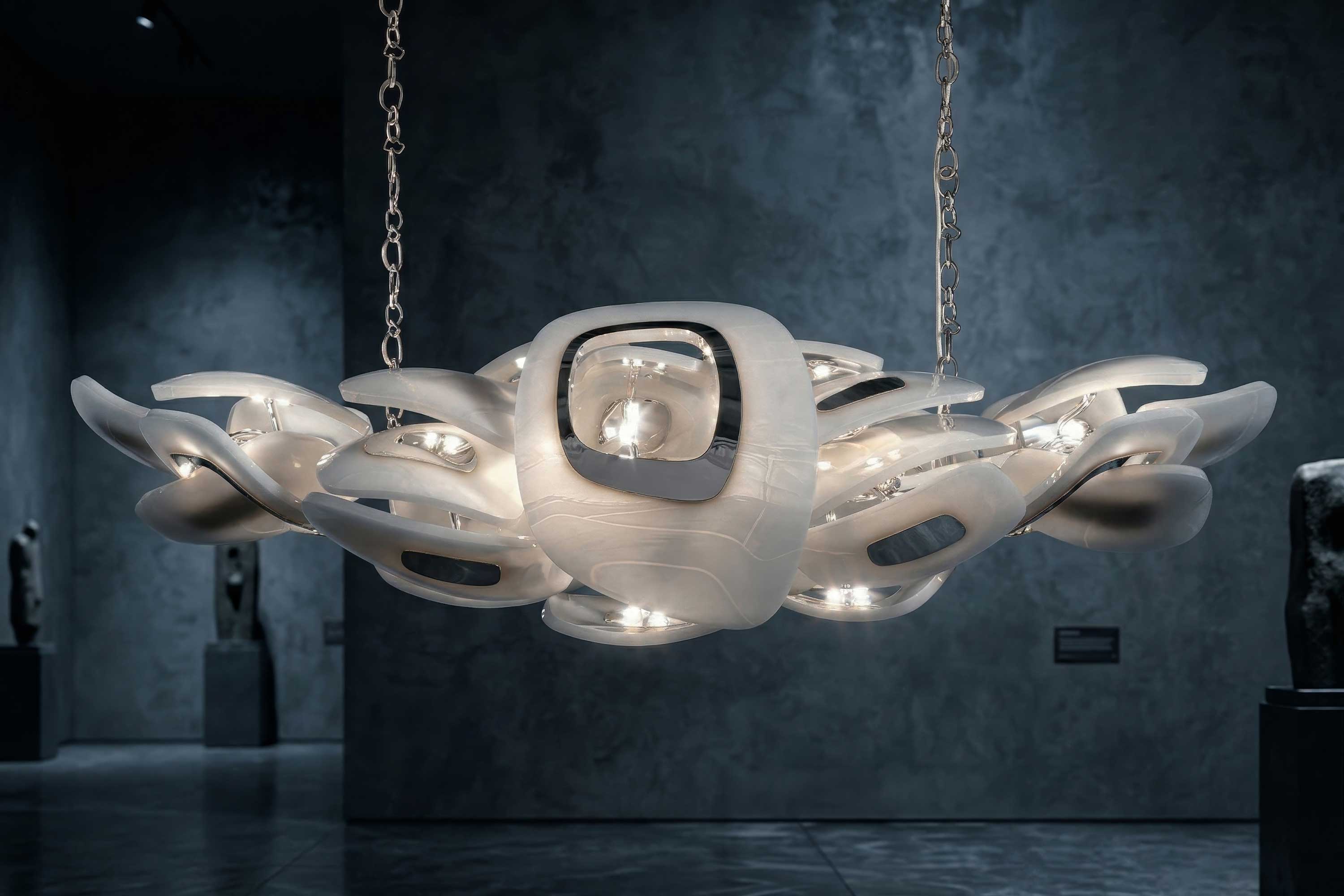 A modern chandelier with interlocking, curved translucent panels is suspended from the ceiling by chains. The chandelier emits soft white light in a dim, gray-walled room with sculptures visible in the background.