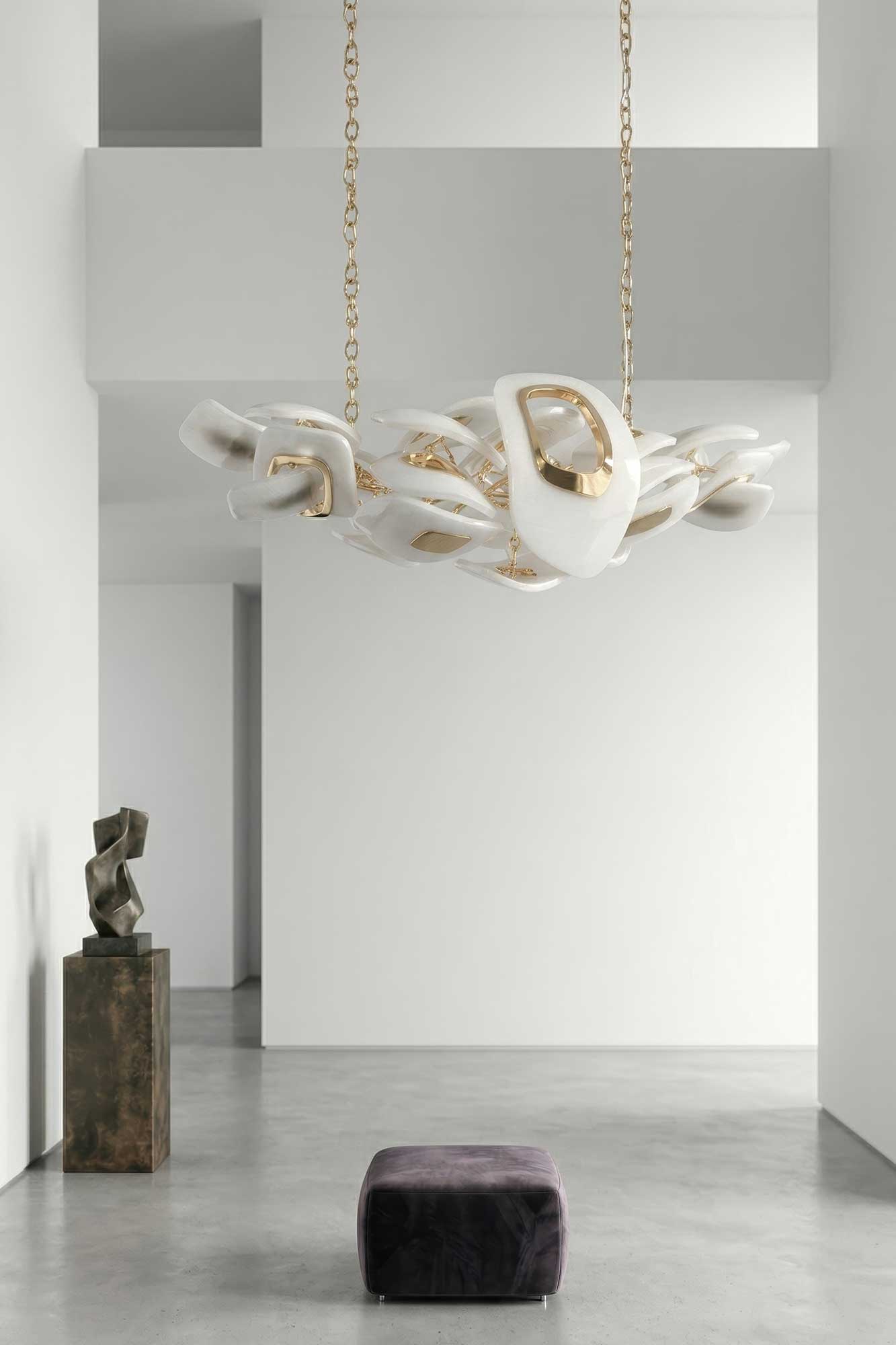A modern white and gold chandelier hangs from the ceiling of a minimalist room with white walls, a dark pedestal sculpture, and a small purple ottoman on a polished concrete floor.