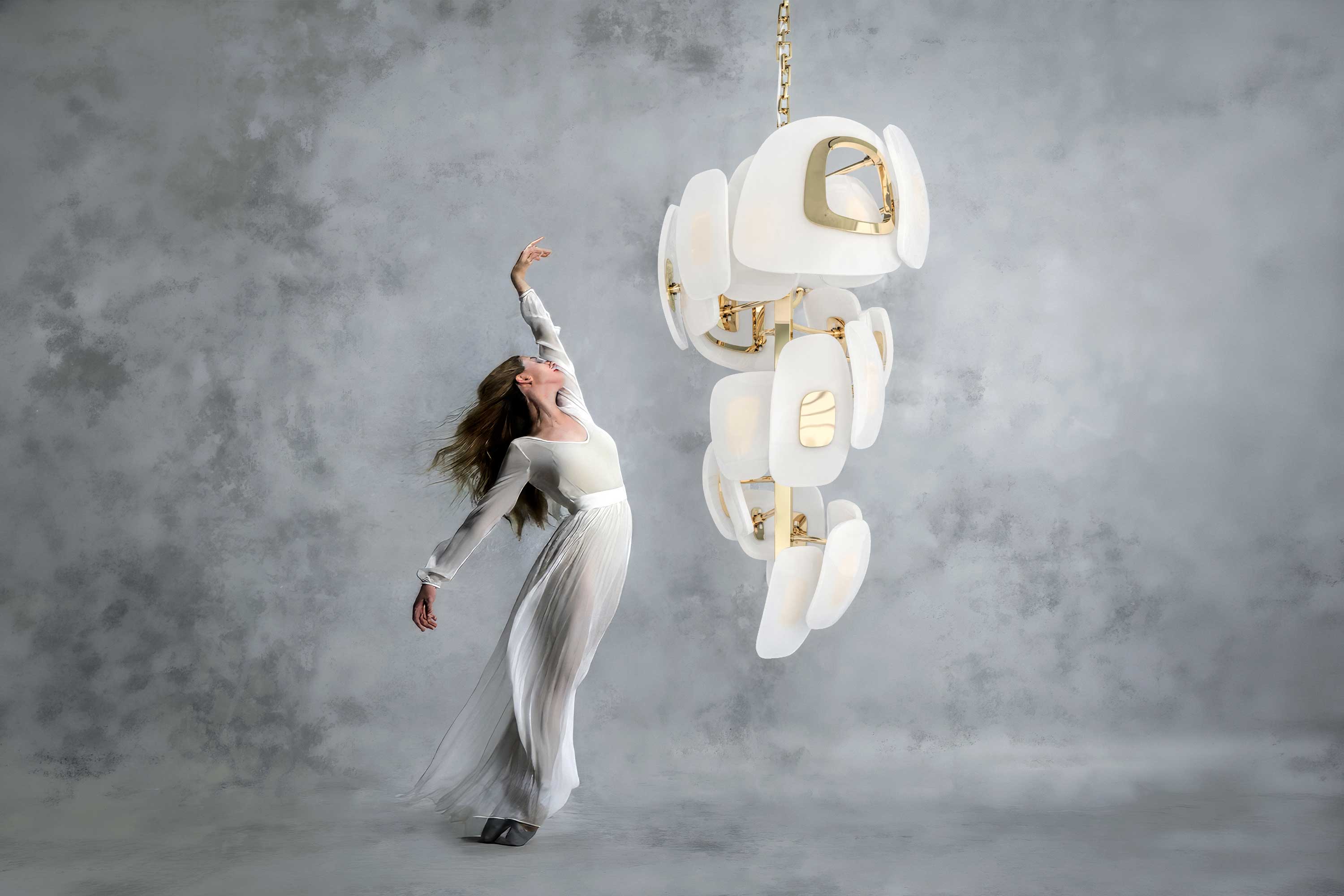 A woman in a long, white dress poses gracefully on one foot next to a modern white and gold chandelier against a gray, textured background.
