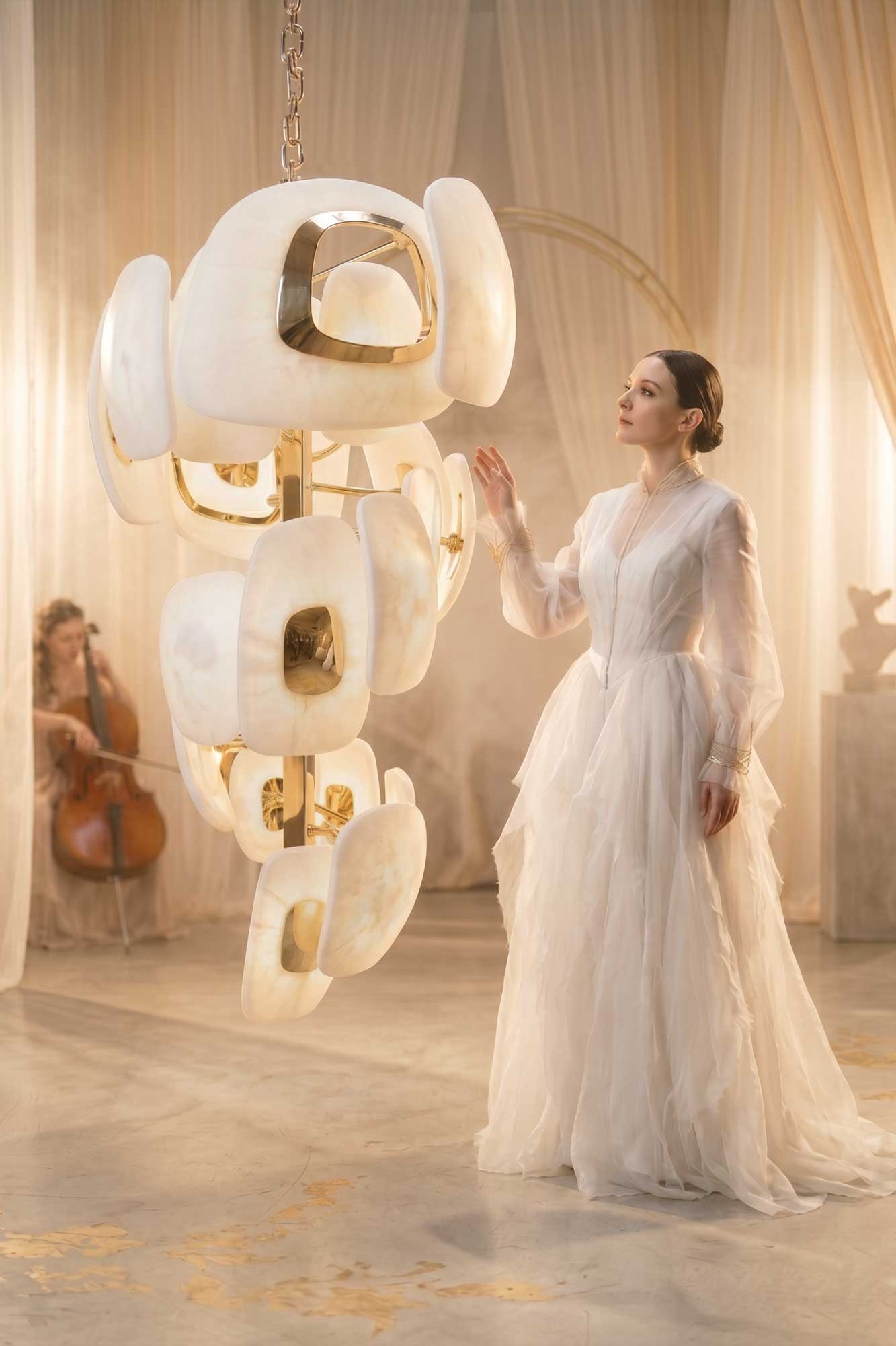 A woman in a flowing white dress stands beside a large, abstract, white and gold hanging sculpture. A musician playing cello is seen seated in the background. The setting is softly lit with sheer curtains.