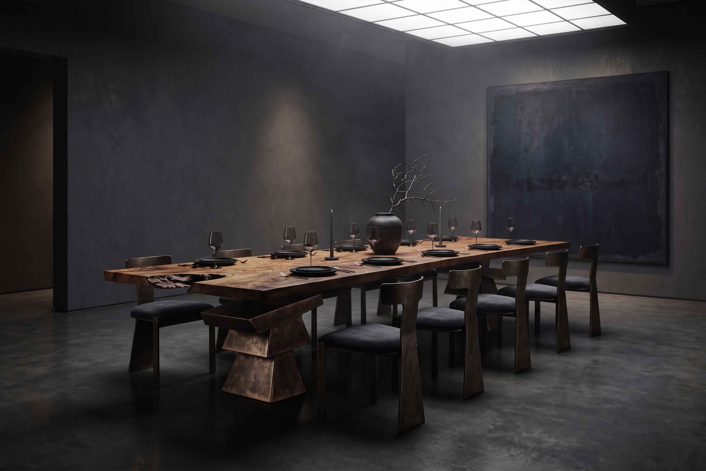 A large wooden dining table with eight dark chairs, set with black plates, glasses, and cutlery. A vase with bare branches sits in the center. The room has dark walls, concrete floors, and minimal lighting.