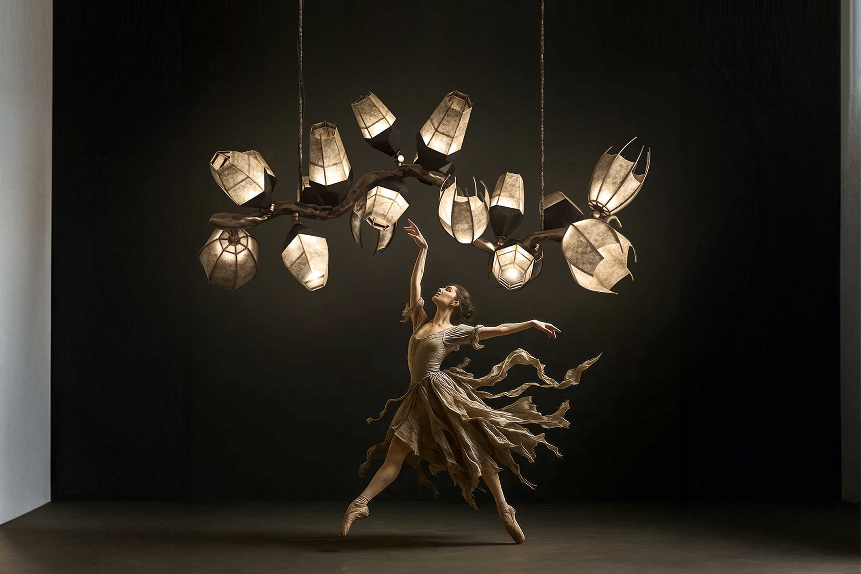 A ballet dancer in a flowing brown dress poses on pointe beneath a large, artistic chandelier resembling tree branches and glowing leaves against a dark background.