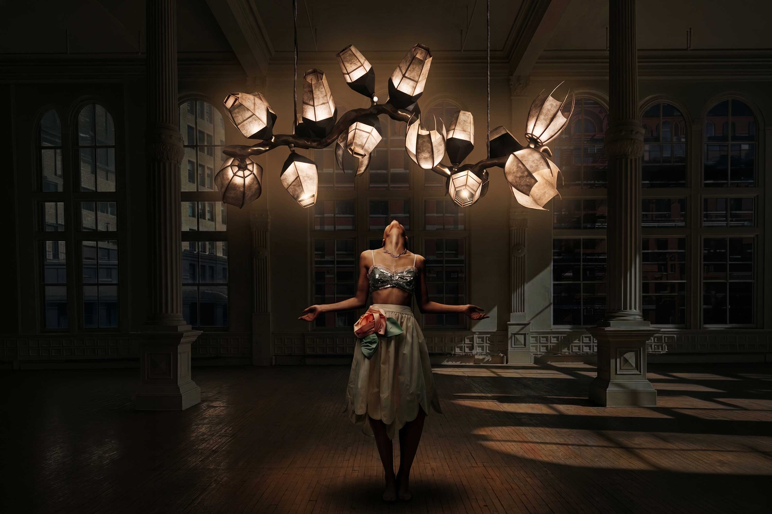 A woman in a skirt and crop top stands in a large, dimly lit room with tall windows, looking up at an abstract, modern chandelier hanging above her. Light from the fixture illuminates her and part of the wooden floor.