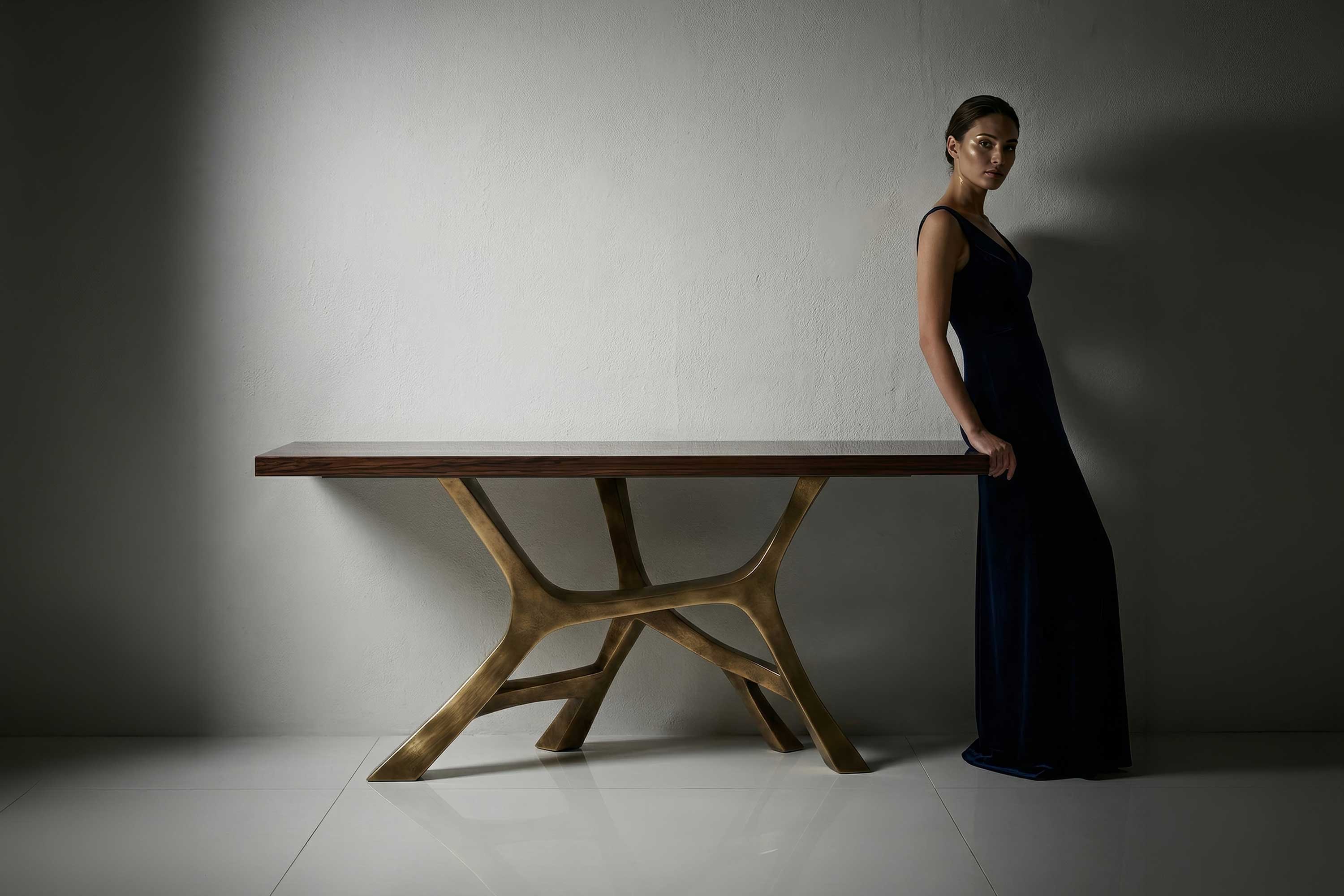A woman in a long, dark dress leans against a modern wooden table with an artistic base, positioned against a plain white wall and white floor.