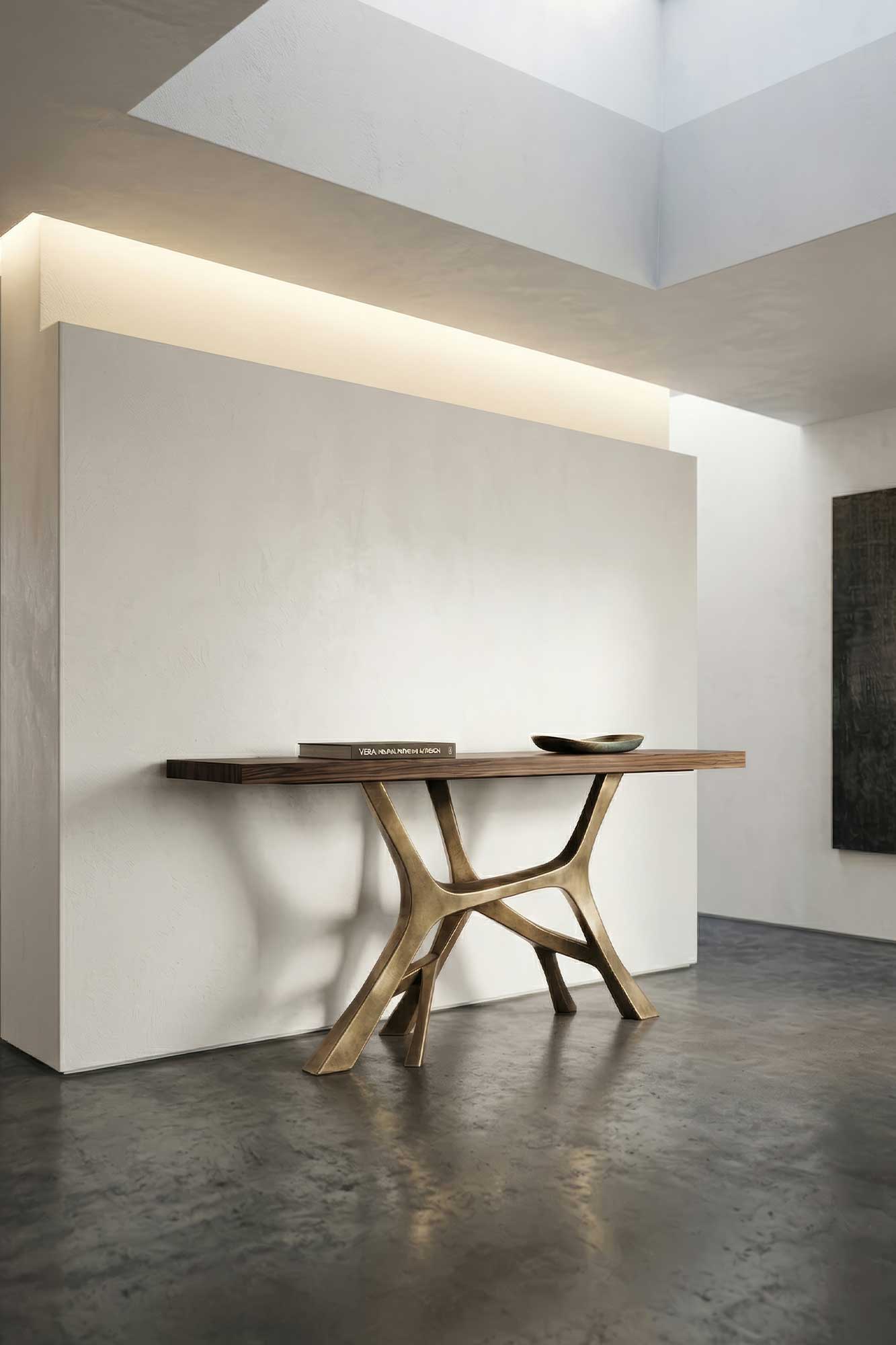 A modern console table with a sculptural bronze base and a wooden top stands against a white wall in a minimalist room. A shallow bowl and a closed book are placed on the table. Light enters from a ceiling window.