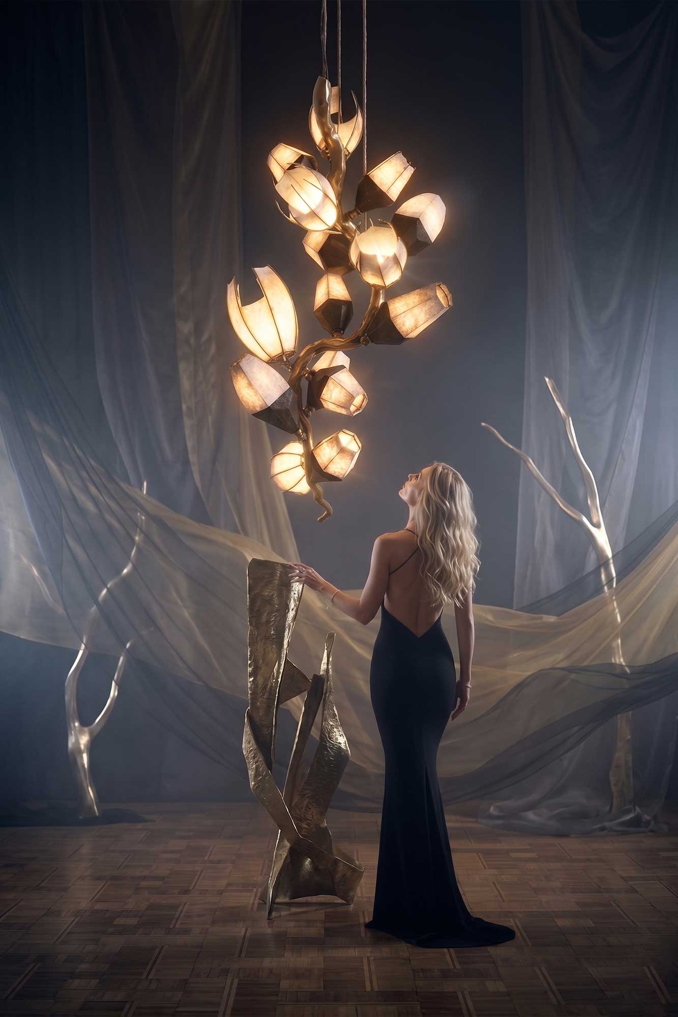 A woman in a long black dress stands on a wooden floor, looking up at a sculptural, modern light fixture. She is surrounded by abstract golden sculptures and draped translucent fabric in a dimly lit room.