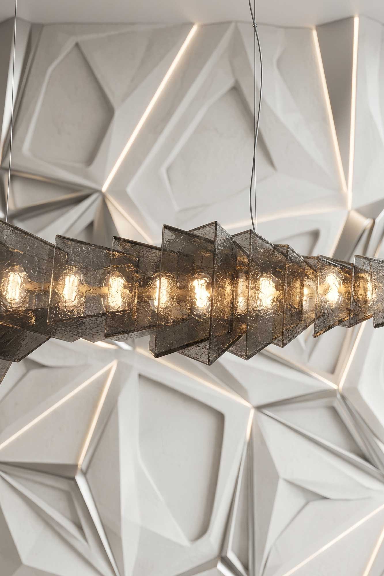 Gellus chandelier in frosted cast glass with a polished bronze core, close view of smoke toned panels lit against a faceted white wall — Palena Studio